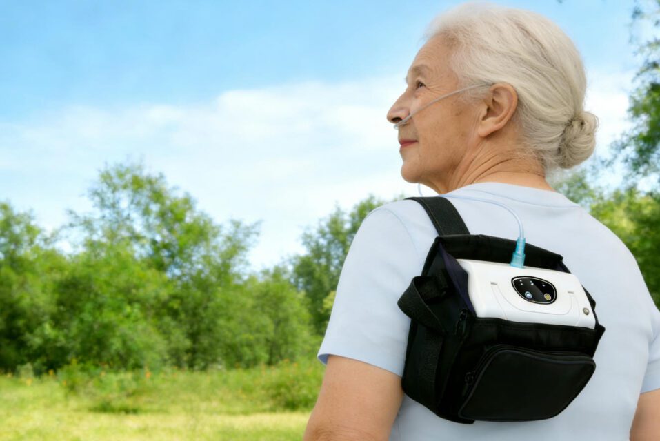 Senior Adult Using Portable Oxygen Concentrator Outdoors - Elderly woman with nasal cannula wearing a backpack carry bag for the device, enjoying nature in a grassy, tree-lined area